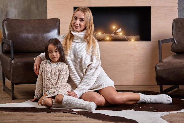 Mãe com menina relaxando no chão perto de um sofá aconchegante com luzes de natal na lareira