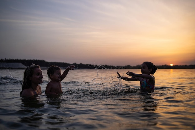 Mãe com o bebê que ela segura em seus braços, brincando com sua filha e espirrando no lago contra o pôr do sol