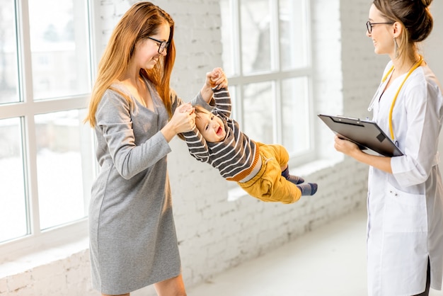 Mãe com seu filho durante a consulta com uma jovem pediatra brincando no escritório branco