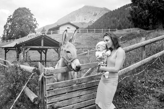 Mãe com sua filha se divertindo no rancho da fazenda e conhecendo um burro Conceito de terapia de animais de estimação no campo com burro na fazenda educacional Conceito de terapia de animais de estimação com crianças Preto e branco