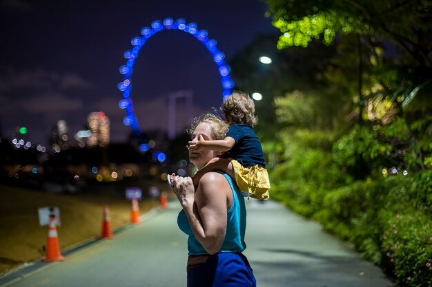 mãe com um bebê nos ombros no parque da cidade à noite contra o fundo da roda gigante