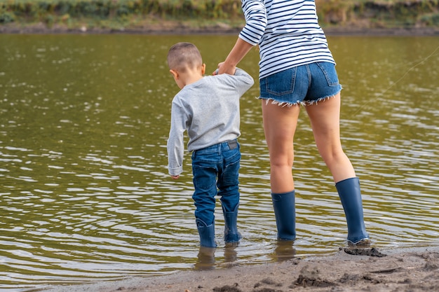 Mãe com um filho pequeno caminha ao longo da costa arenosa do lago em botas de borracha