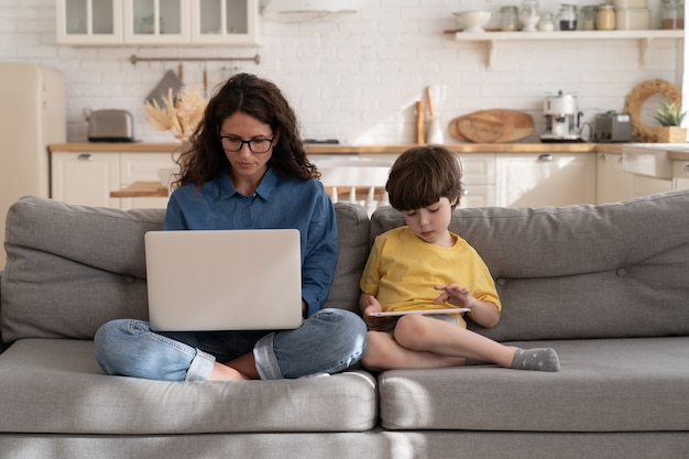 Mãe concentrada, filho, usa laptop e tablets, senta no sofá da sala de estar, trabalha e estuda