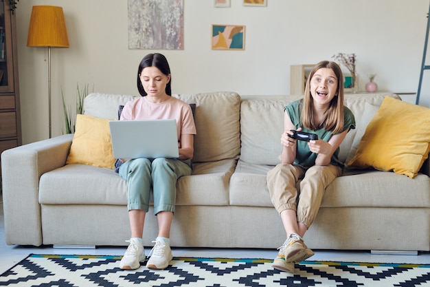 Mãe concentrada usando laptop e trabalhando em casa enquanto a filha joga videogame