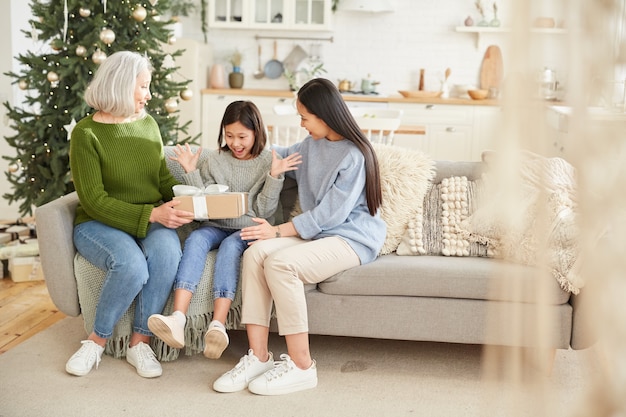 Mãe dando um presente para sua filha mais nova no dia de Natal, enquanto elas estão sentadas no sofá da sala de estar