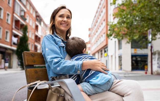 Mãe de 40 anos abraçando seu filho de dois anos na rua em um dia nublado feliz sentado em um banco