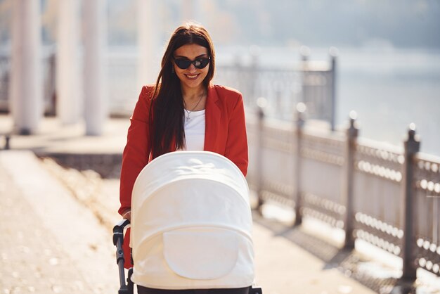 Mãe de casaco vermelho dá um passeio com seu filho no carrinho de bebê no parque no outono.