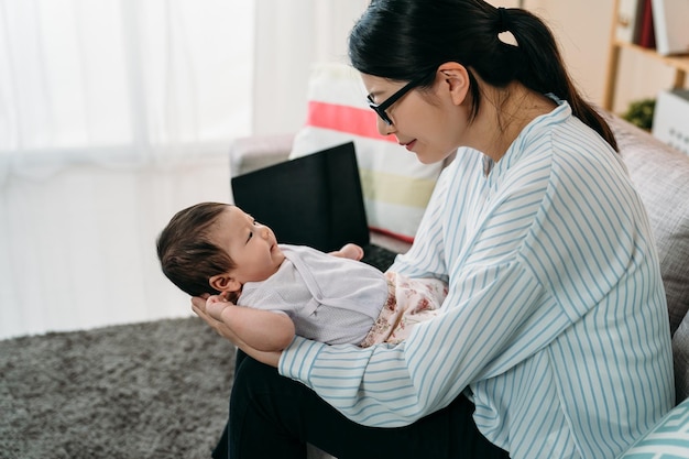 Mãe de empresária asiática de retrato lateral está segurando e olhando para sua filha bebê fofo amorosamente em uma sala de estar brilhante em casa.