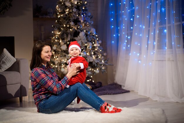 Mãe de família feliz e bebê perto de árvore de Natal à noite de feriado.