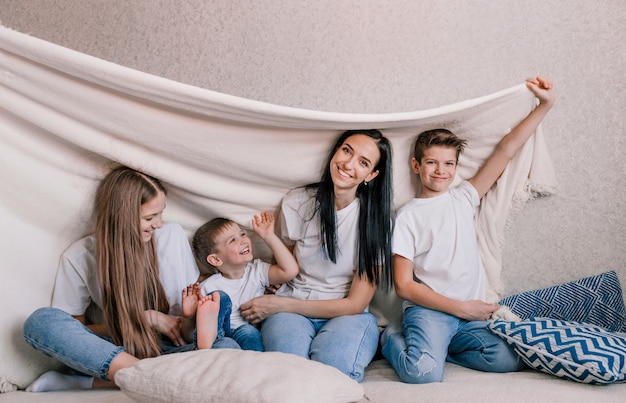 Mãe de família feliz e crianças pequenas riem alegremente sentados no sofá coberto com um cobertor branco. felicidade familiar fim de semana divertido