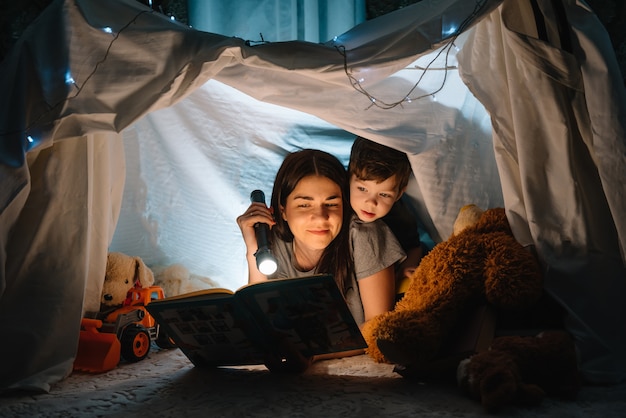 Mãe de família feliz e filho lendo um livro com uma lanterna em uma tenda em casa. conceito de familia