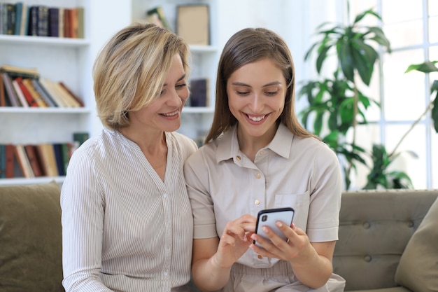 Mãe de meia-idade e filha adulta se abraçando, usando o telefone juntas, assistindo a vídeos ou fotos, sentadas no sofá aconchegante em casa.