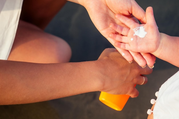 Mãe de óculos elegantes e vestido branco está colocando o frasco de spray laranja SPF no bebê recém-nascido, filha de um ano, um protetor solar na praia