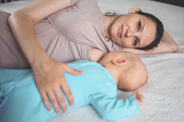 Mãe deita-se com bebê recém-nascido em macacão azul abraçando e amamentando com leite materno