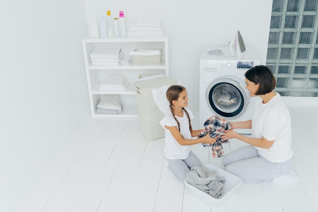 Mãe dona de casa e filha carregam máquina de lavar com roupas sujas posam na espaçosa lavanderia faz tarefas em casa olham alegremente uma para a outra Conceito de dia da lavanderia Família na lavanderia
