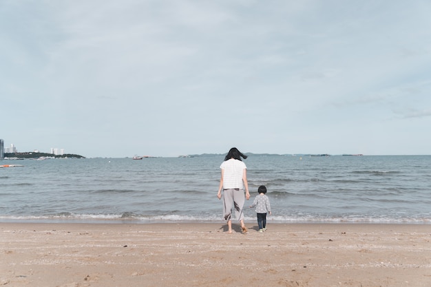 Mãe e criança asiática de 3 anos caminhando na praia.