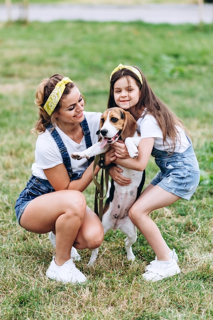 Mãe e filha, abraçando e brincando com o cachorro ao ar livre.