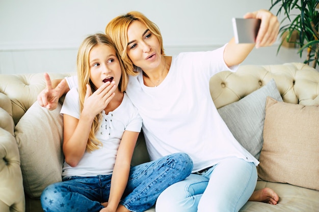 Mãe e filha abraçando, posando e fazendo selfie no telefone inteligente enquanto está sentado no sofá em casa