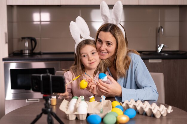 mãe e filha caucasianas com um curativo nas orelhas de um coelho estão sentadas na cozinha em casa decorando ovos de Páscoa tirando uma selfie usando um smartphone e um tripé