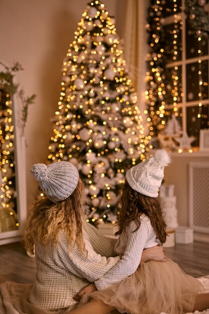 Mãe e filha com chapéus de malha olhando para a árvore de Natal brilhante