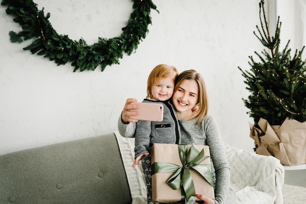 Mãe e filha com presentes são fotografadas no telefone perto da árvore de Natal Mãe e filho se divertindo abraçando e brincando juntos perto de casa Feliz Natal e Boas Festas