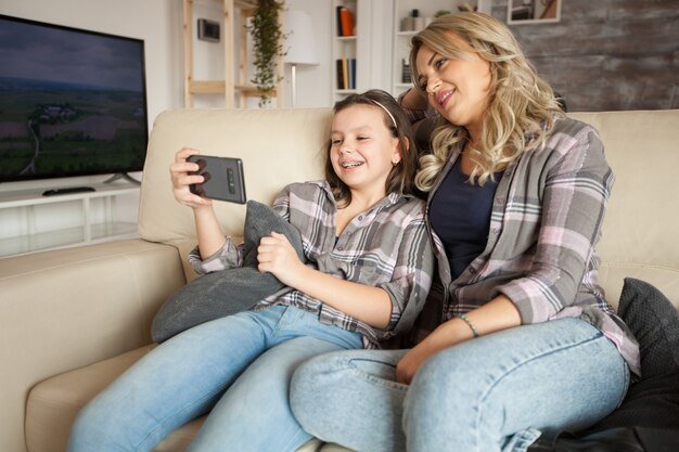 Foto mãe e filha com roupas combinando, sentadas no sofá assistindo a um vídeo engraçado no smartphone.