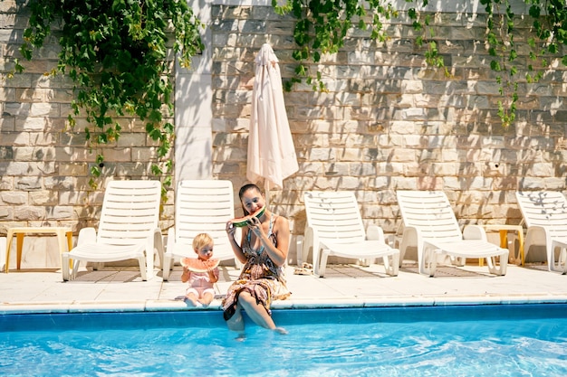 Mãe e filha comem melancia sentada à beira da piscina