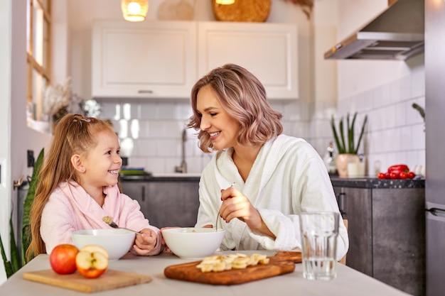 Mãe e filha comendo frutas e mingau. nutrição saudável para crianças, refeição matinal. Família caucasiana tomando café da manhã em uma cozinha moderna e leve