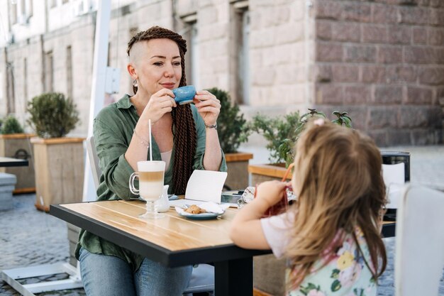 Mãe e filha comendo no café de rua moderna mãe hipster com dreadlocks e