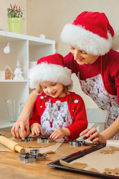 Mãe e filha cortam biscoitos de gengibre de Natal para as boas festas em chapéus de Papai Noel