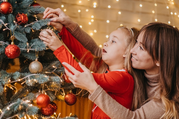 Mãe e filha decoram a árvore de Natal juntas. Paternidade tradicional.