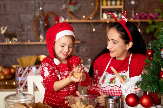 Mãe e filha em uma cozinha escura com uma árvore de natal cozinham biscoitos de gengibre no ano novo ou no natal e sorriem com um chapéu de papai noel