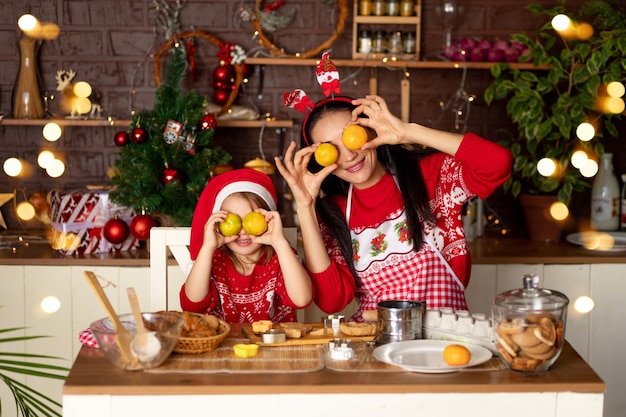 Mãe e filha em uma cozinha escura com uma árvore de Natal estão preparando biscoitos de gengibre para o Ano Novo ou o Natal, sorrindo e brincando com tangerinas em um chapéu de Papai Noel