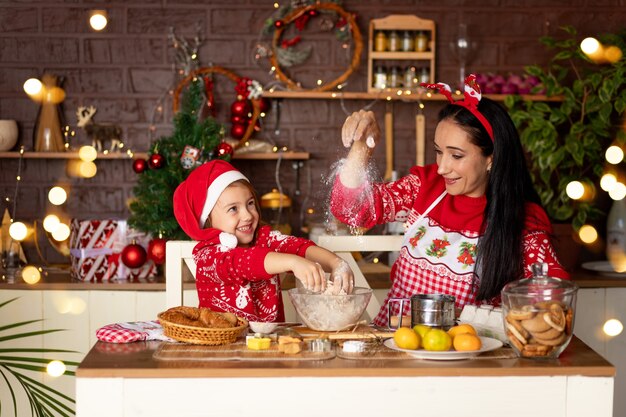 Mãe e filha em uma cozinha escura com uma árvore de Natal estão preparando biscoitos de gengibre para o Ano Novo ou o Natal, sorrindo e se divertindo juntas em antecipação ao feriado com um chapéu de Papai Noel