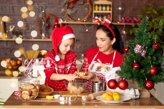 Mãe e filha estão cozinhando biscoitos de gengibre na cozinha e se preparando para o ano novo e o Natal, se divertindo e se alegrando com suéteres vermelhos e chapéu de Papai Noel