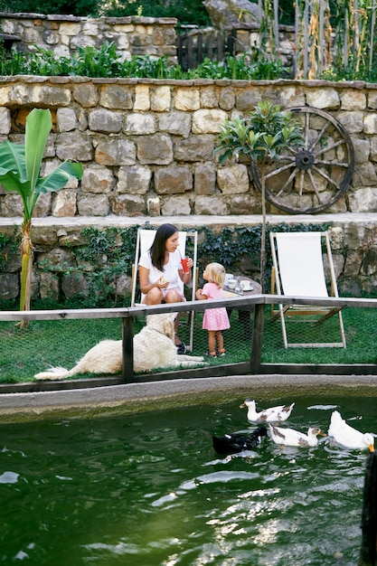 Mãe e filha estão sentadas a uma mesa no gramado perto do lago com patos