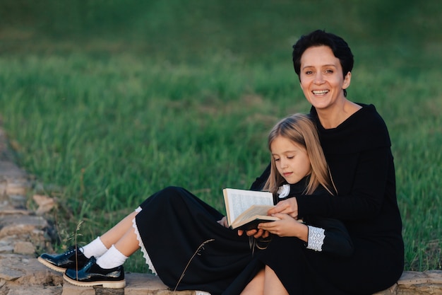 Foto mãe e filha estão sentadas em um banco de pedra e lendo um livro. mulher com uma criança em vestidos pretos.