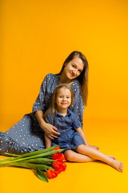 Mãe e filha estão sentadas no chão com um buquê de tulipas vermelhas e olham para a frente em uma parede amarela