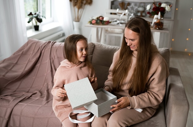 Mãe e filha estão sentadas no sofá com uma caixa de presente e abrindo-a