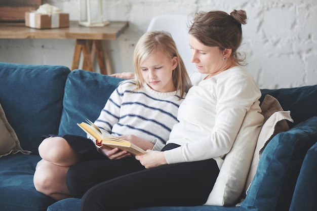 Mãe e filha estão sentadas no sofá do apartamento. menina lendo