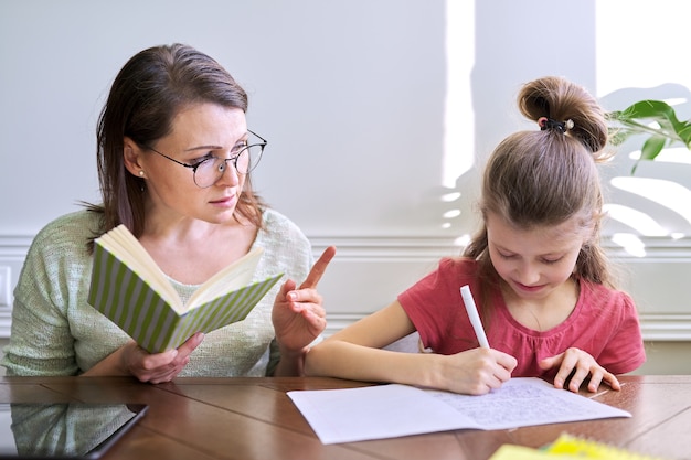 Mãe e filha estudam juntos em casa, sentados à mesa. Mulher com livro, garota escreve no caderno. Aprendizagem à distância, pais ajudando aluno da escola primária