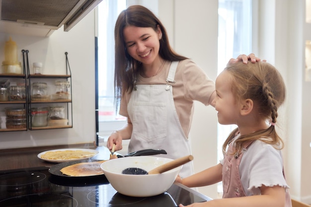 Mãe e filha fritaram panquecas juntas na cozinha em um brilhante interior, assistente de mães e brea