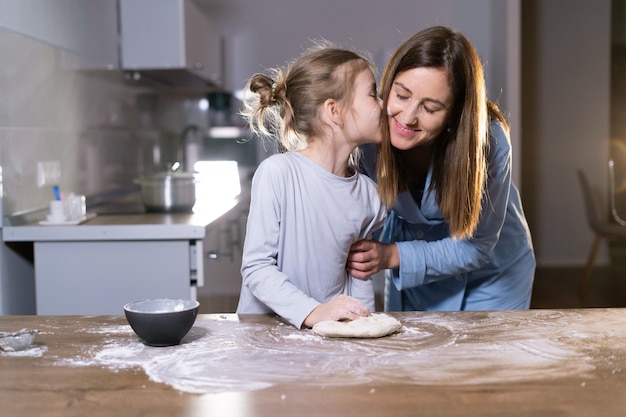 Mãe e filha juntas na cozinha amassam a massa a garota beija a mulher conceito de ajuda para pais Copiar espaço