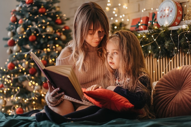 Mãe e filha lendo contos de fadas na véspera de Natal, atmosfera maravilhosa de amor e confiança, crença em milagres, boas festas