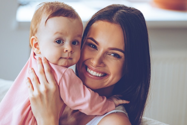 Foto mãe e filha. mulher jovem e bonita alegre segurando a menina nas mãos e olhando para a câmera com um sorriso enquanto está sentado no sofá em casa