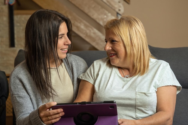 Mãe e filha no sofá em casa enquanto se olham com cumplicidade e segurando um tablet e sendo felizes