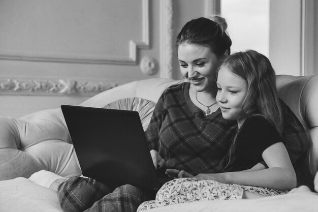 Mãe e filha passam um tempo juntos com o laptop em casa, na sala de estar no sofá. Feliz mãe e filha caucasiana usando computador, rindo, navegando na Internet, fazendo compras online, procurando um filme