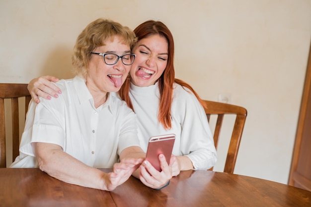 Mãe e filha são fotografadas na câmera frontal do smartphone enquanto estão sentadas em casa à mesa Alegre mãe idosa e sua filha fazendo selfies cômicas e mostrando línguas