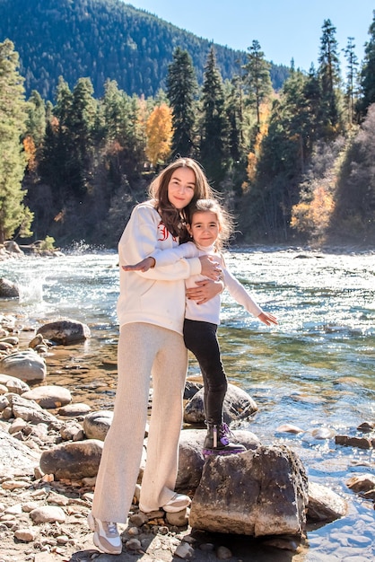 Mãe e filha se abraçam no cenário de um rio de montanha alpina com pinheiros nas colinas