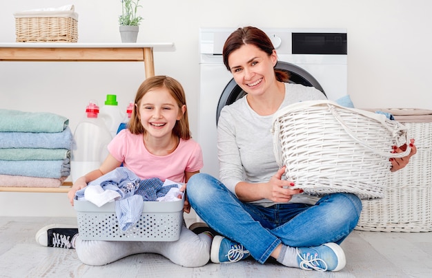 Mãe e filha se divertindo durante a lavanderia sentadas no chão com cestos de roupa na sala iluminada